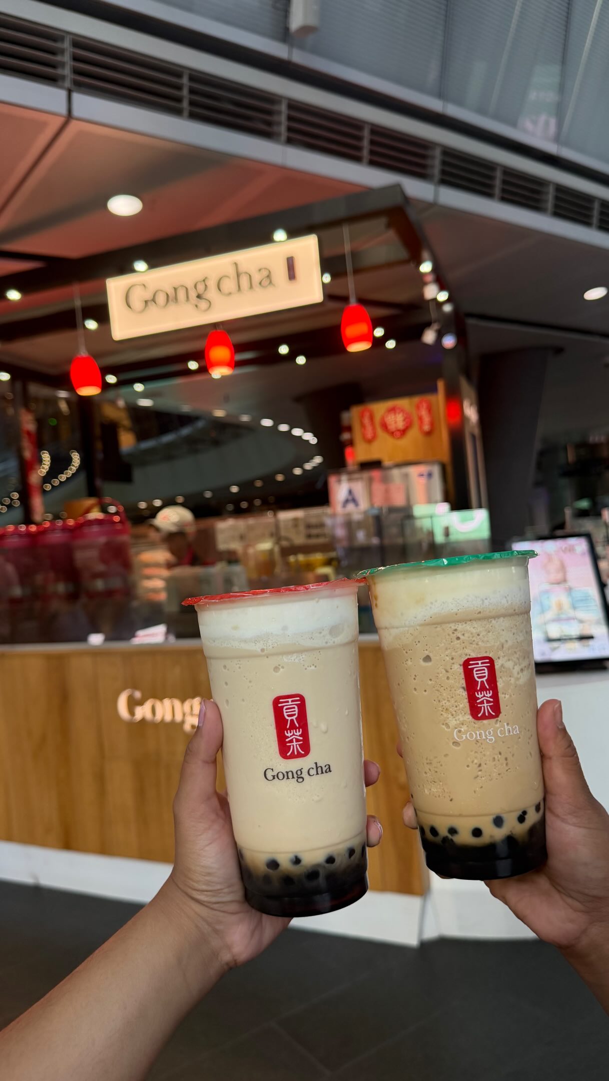Two hands holding Gong cha drinks in front of a Gong cha storefront